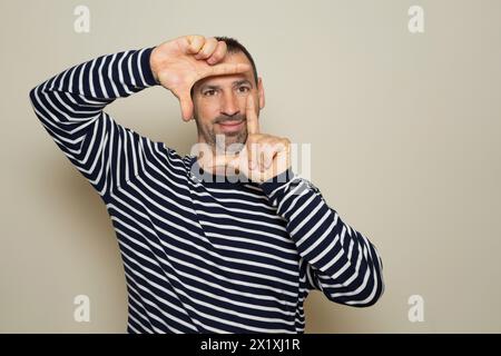 Un uomo barbuto felice sbatte gli occhi e guarda attraverso una cornice formata dalle sue mani. Fa una cornice con le dita, isolata su sfondo beige Foto Stock