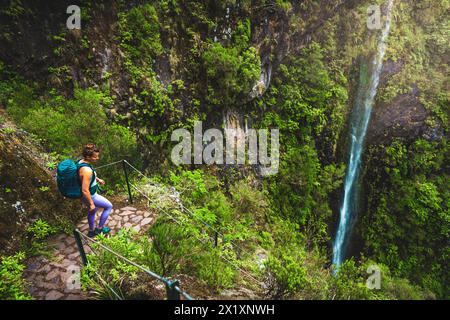 Descrizione: Una donna turista in viaggio con zaino in spalla gode di una vista sulla cascata ricoperta mentre percorre un sentiero simile a una scala nella foresta pluviale. Levada di Caldeirão Verd Foto Stock