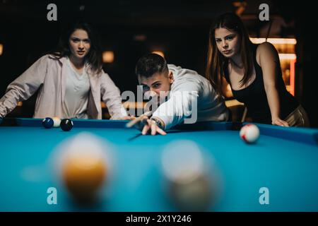 Uomo concentrato che gioca a biliardo con amiche in un bar poco illuminato Foto Stock