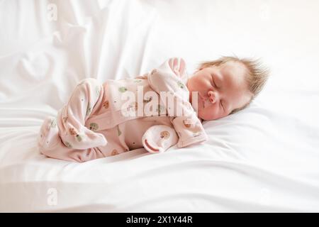 Piccolo neonato caucasico che dorme su biancheria da letto bianca indossando una tuta rosa. Neonata che dorme Foto Stock