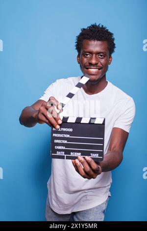 Il regista afroamericano tiene in mano il clapper aperto e si sente sicuro di sé con un sorriso sul viso. Produttore cinematografico nero con lavagna in legno di legno di fronte a sfondo blu isolato. Foto Stock