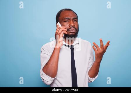 Un freelancer maschio che sta cercando di spiegare il problema dello smartphone che fa il gesto della mano su sfondo blu. Turbato uomo d'affari afroamericano di cattivo umore che cerca di capire la conversazione d'affari. Foto Stock
