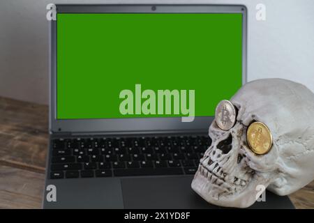 laptop cromakey con schermo verde mockup, modello di cranio umano sulla tastiera, bitcoin metallici nelle prese oculari, concetto di estrazione di criptovalute, Foto Stock