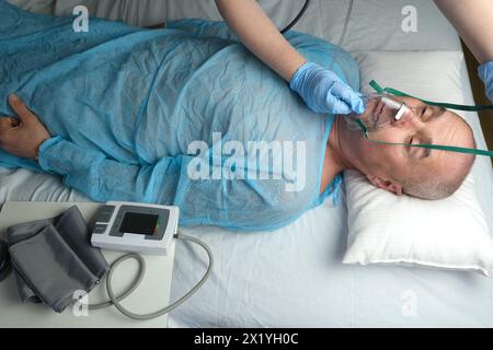 uomo anziano paziente in camicia blu bugie, il medico mette su maschera di ossigeno facciale, concetto di assistenza medica, anestesiologia e terapia intensiva, ossigeno ther Foto Stock