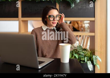 Donna che indossa gli occhiali con un notebook. Affascinante donna d'affari con occhiali da vista e abiti casual che lavora con un computer portatile mentre si trova in una caffetteria. Elegante donna che regola gli occhiali guardando il lato Foto Stock