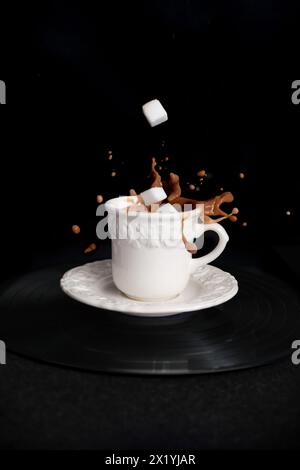 primo piano del caffè cappuccino in una tazza bianca e piattino su sfondo nero con schizzi e gocce di liquido in tutte le direzioni, gocce di zucchero raffinate, Foto Stock