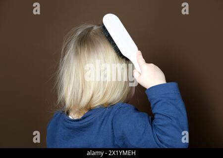 bambina bionda che pettina i capelli in modo indipendente con una spazzola di legno con setole naturali, bambina su sfondo marrone scuro, concetto di igiene, hai Foto Stock