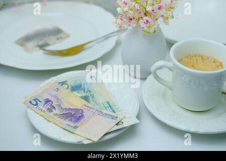 Vista dall'alto primo piano su piatto bianco con banconote e monete di yuan cinesi, attenzione selettiva, tazza di caffè, piatti con cibo, pranzo al ristorante, caffè Foto Stock