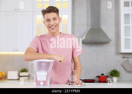 Uomo felice con caraffa filtro e bicchiere d'acqua pulita a tavola in cucina Foto Stock