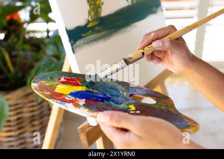 Una giovane donna birazziale dipinge su tela a casa, mescolando colori su una tavolozza Foto Stock