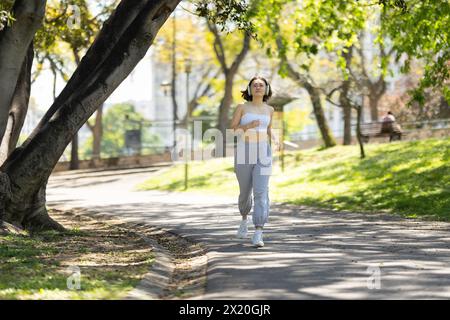 Una donna corre su un sentiero in un parco. Indossa le cuffie e una canotta bianca Foto Stock