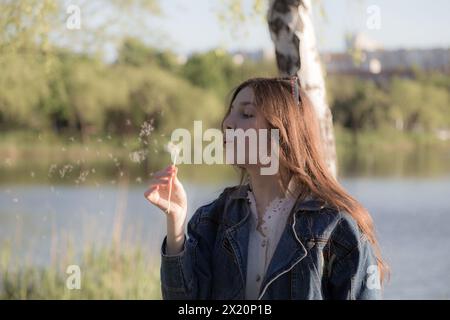 Una bella ragazza soffia su un dente di leone. Sta succedendo da qualche parte nel parco. Primo piano. Foto Stock