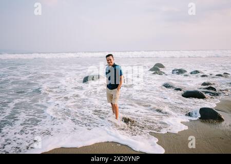 Un uomo soddisfatto con una maglietta blu e pantaloncini si trova sulla riva, il suo sguardo fisso verso l'orizzonte, irradiando felicità sullo sfondo del beac Foto Stock