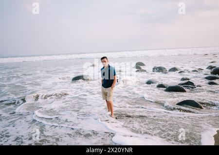 Un uomo soddisfatto con una maglietta blu e pantaloncini si trova sulla riva, il suo sguardo fisso verso l'orizzonte, irradiando felicità sullo sfondo del beac Foto Stock