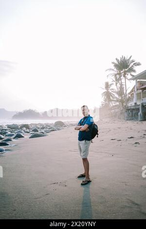 Un uomo soddisfatto con una maglietta blu e pantaloncini si trova sulla riva, il suo sguardo fisso verso l'orizzonte, irradiando felicità sullo sfondo del beac Foto Stock