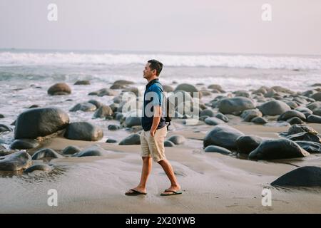 Un uomo soddisfatto con una maglietta blu e pantaloncini si trova sulla riva, il suo sguardo fisso verso l'orizzonte, irradiando felicità sullo sfondo del beac Foto Stock