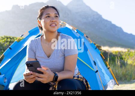 Escursionista birazziale con smartphone, seduto accanto alla tenda Foto Stock