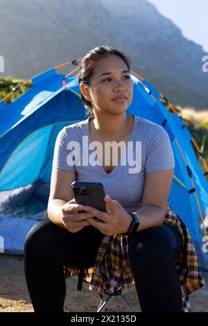 Escursionista birazziale con smartphone, seduto accanto alla tenda Foto Stock