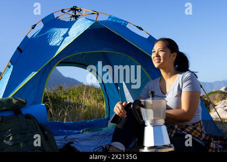 Escursionista birazziale seduto accanto alla tenda, tenendo la caffettiera, guardando lontano Foto Stock