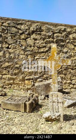 Un muro di pietra con una croce davanti. La croce è vecchia e ha un muschio giallastro. Antico cimitero Foto Stock