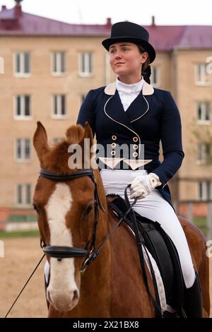 Dressage cavaliere su castagno con sfondo urbano. Sessione di guida. Fantino femminile in equino uniforme. Sport equestre. Equitazione schoo Foto Stock