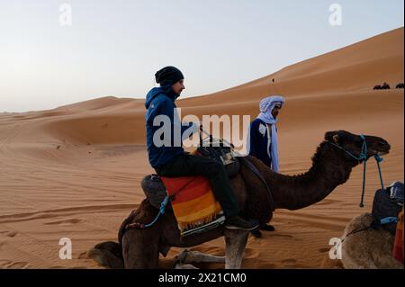 Un uomo di mezza età si prepara a scendere a bordo di un cammello, aiutato da un addetto locale mentre l'animale si inginocchia, con una duna di sabbia che si innalza nel deserto marocchino. Foto Stock