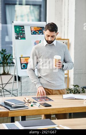 Un uomo si rilassa a un tavolo con una tazza di caffè in un vivace ambiente d'ufficio, prendendosi un momento per riorganizzarsi nel caos aziendale. Foto Stock