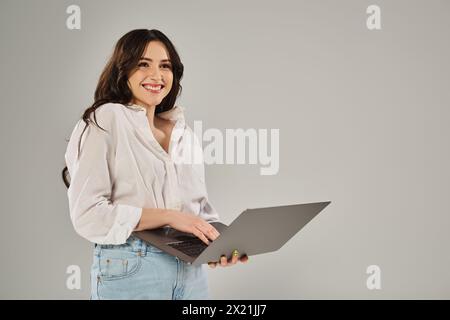 Una donna elegante e di grandi dimensioni sorride tenendo un computer portatile su uno sfondo grigio. Foto Stock