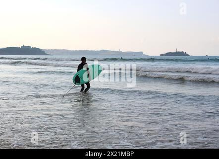 Un surfista che cammina nella spiaggia di Somo con il faro di Mouro Island El Faro e la penisola di Magdalena Ribamontán al Mar Cantabria Spagna Foto Stock