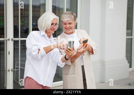 Due gioiose donne anziane in piedi fuori dall'edificio, sorridenti scambiano i dati di contatto con gli smartphone, indossano un elegante abbigliamento casual e si godono il calore Foto Stock