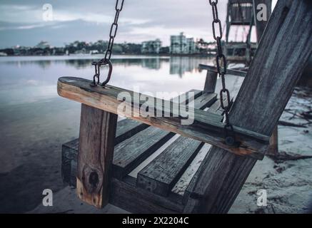 Un'altalena di legno intemprata è appesa a catene su una spiaggia di legno. Il fascino rustico dell'altalena contrasta con gli edifici moderni sullo sfondo. Foto Stock
