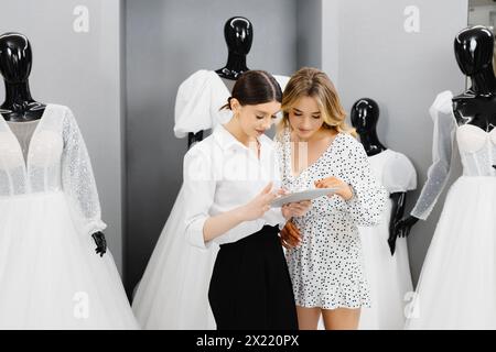 Splendida sposa che parla di dettagli con il wedding planner in negozio. Le attraenti donne proprietarie di negozi nuziali utilizzano un tablet digitale per aiutare i clienti a scegliere il matrimonio Foto Stock