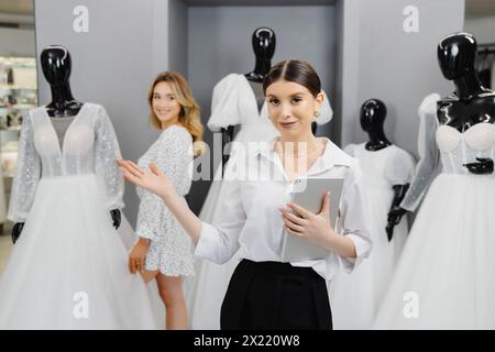 Le attraenti donne proprietarie di negozi nuziali utilizzano un tablet digitale per aiutare i clienti a scegliere l'abito da sposa nel negozio. Foto Stock