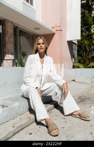 Una giovane donna serena dai capelli biondi, elegantemente vestita con un abito bianco, si siede graziosamente a Miami. Foto Stock