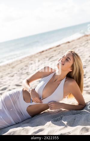 Una giovane e bella donna bionda si stende pacificamente sulla spiaggia sabbiosa di Miami Beach, immergendosi nel caldo abbraccio del sole. Foto Stock