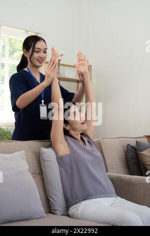 Donna anziana felice che fa esercizio fisico a casa con fisioterapista asiatica. Una vecchia signora in pensione che fa stretching le braccia a casa con l'aiuto di un Foto Stock