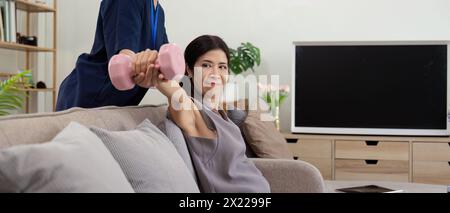 Una donna anziana che si allena con fisioterapista usando i manubri a casa. Terapeuta asiatica che assiste una donna anziana nell'esercizio fisico in casa di riposo. Paziente anziano Foto Stock