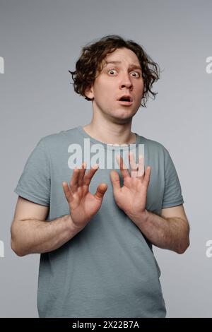 Ritratto verticale medio in studio di un giovane uomo caucasico con capelli ondulati che dimostrano una sensazione di paura, sfondo grigio Foto Stock