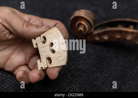 Mani di un liutaio latinoamericano irriconoscibile che regge un ponte per violino, sullo sfondo una testa di violino. Concetto di strumenti a corda Foto Stock