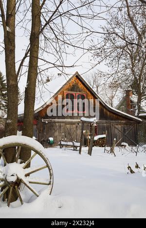 Vecchio fienile rustico in legno con finestre rifinite rosse incorniciate da alberi nel giardino sul retro in inverno, Quebec, Canada Foto Stock