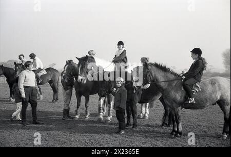 Anni '1970, storica, fuori in un campo, in un centro di equitazione, un certo numero di ragazze sedute su cavalli che hanno una lezione di equitazione, con istruttore maschio adulto, Inghilterra, Regno Unito. In questa lezione, i giovani imparano le basi di un cavallo, tra cui come usare le gambe e le mani in un posto lento, che conduce a un trotto e poi con buon equilibrio e ritmo, fino alla fase successiva che è una cantera. Foto Stock