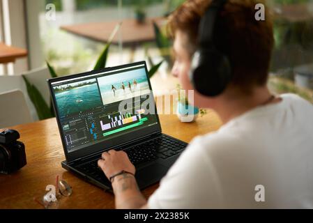L'editor video professionale con cuffie funziona su un computer portatile in un bar. Vista a spalla, schermata software di editing, timeline colorata. Macchina fotografica, occhiali sul tavolo Foto Stock