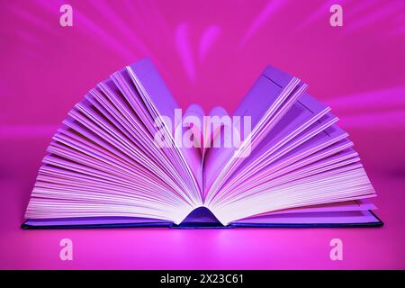 Apri libro su sfondo rosa illuminato dalla parte anteriore con interessanti sequenze di luce dietro e pagine ruotate a forma di cuore Foto Stock