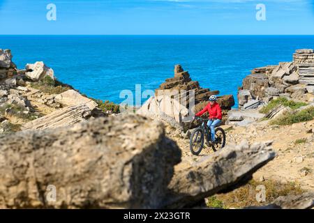 Coraggiosa donna anziana in sella alla sua mountain bike elettrica sulle scogliere rocciose di Peniche, sulla costa atlantica occidentale del Portogallo Foto Stock