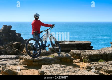 Coraggiosa donna anziana in sella alla sua mountain bike elettrica sulle scogliere rocciose di Peniche, sulla costa atlantica occidentale del Portogallo Foto Stock