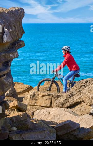 Coraggiosa donna anziana in sella alla sua mountain bike elettrica sulle scogliere rocciose di Peniche, sulla costa atlantica occidentale del Portogallo Foto Stock