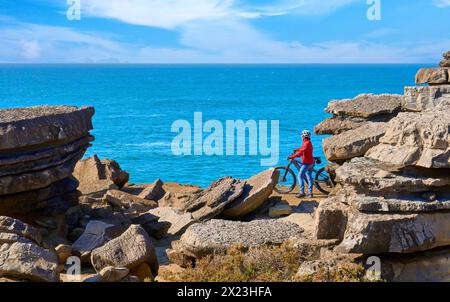 Coraggiosa donna anziana in sella alla sua mountain bike elettrica sulle scogliere rocciose di Peniche, sulla costa atlantica occidentale del Portogallo Foto Stock