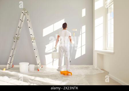 Pittore maschile in abiti bianchi in piedi in una stanza vuota con un secchio e un rullo per pittura Foto Stock