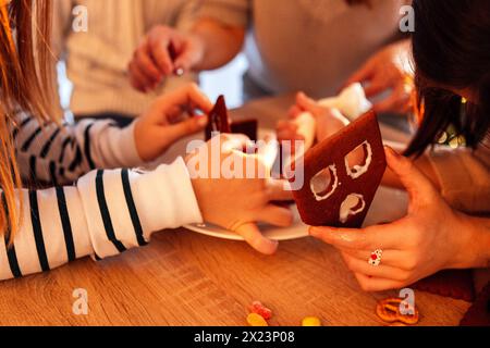 Primo piano di mani femminili che assemblano una casa di pan di zenzero su un tavolo di legno. Le ragazze decorano i biscotti di Natale con dolci. Cucina festiva con il whol Foto Stock