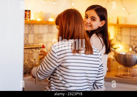 Due ragazze adolescenti adorabili stanno lavando i piatti dopo aver cucinato a casa in cucina. Le sorelle sorridenti stanno facendo i compiti. Amici che passano il tempo e Wo Foto Stock